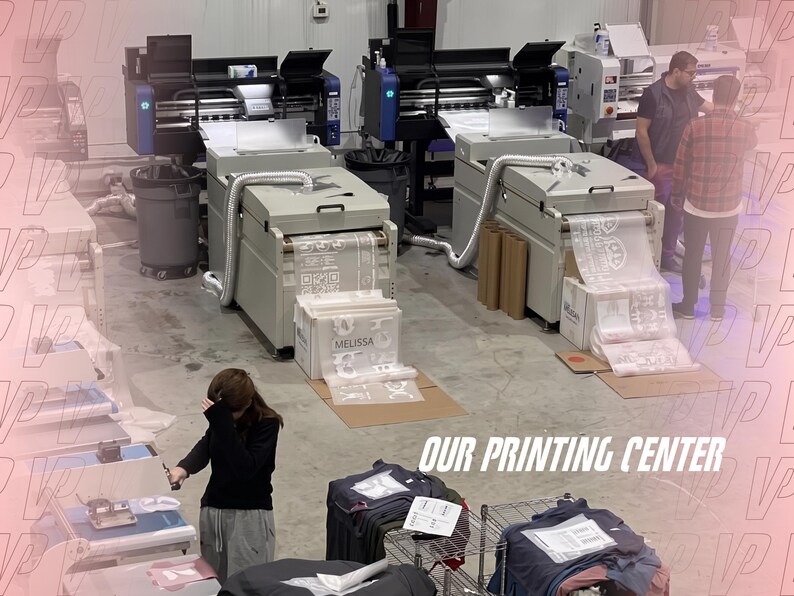 May include: A printing center featuring two large industrial printers with white output sheets, a person standing near the machines, and another person working at a station with stacks of folded garments and the text OUR PRINTING CENTER.