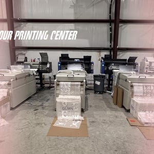 May include: Three industrial printing machines arranged in a row within a facility. Each machine features a light gray housing and is actively processing rolls of white film with printed graphics and text, including the letters GLMN and a QR code.