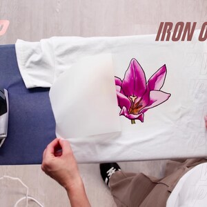 May include: A white t-shirt laid flat on an ironing board with a vibrant pink tulip graphic transfer being applied. A piece of parchment paper partially covers the flower design, and a silver iron sits on the left side of the board.