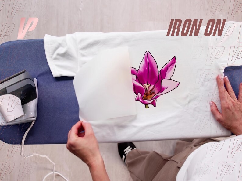 May include: A white t-shirt laid flat on an ironing board with a vibrant pink tulip graphic transfer being applied. A piece of parchment paper partially covers the flower design, and a silver iron sits on the left side of the board.