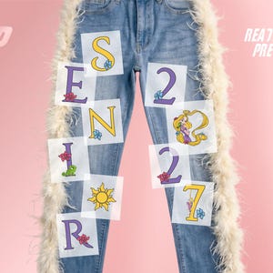 May include: Blue denim jeans featuring white feather trim along the outer seams. The legs are decorated with iron-on transfers spelling SENIOR 2027, incorporating yellow and purple letters, a sun icon, a green lizard, and a cartoon character.