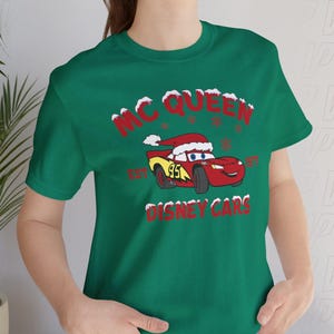 May include: A dark green t-shirt featuring a graphic of the red race car character Lightning McQueen wearing a Santa hat. The text reads MC QUEEN EST 1997 DISNEY CARS, with red lettering and white snow accents surrounding the car and the text.