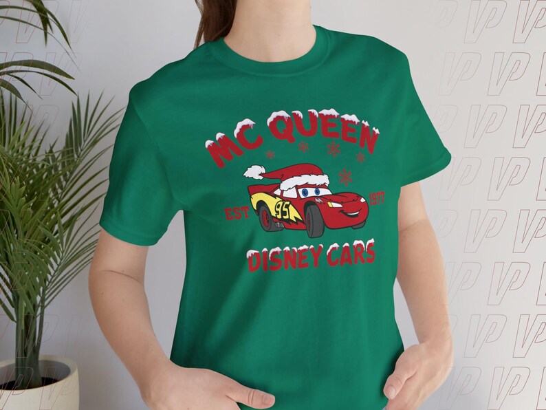 May include: A dark green t-shirt featuring a graphic of the red race car character Lightning McQueen wearing a Santa hat. The text reads MC QUEEN EST 1997 DISNEY CARS, with red lettering and white snow accents surrounding the car and the text.