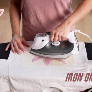 May include: A white electric iron with a dark gray soleplate is being used to press a transfer onto a white t-shirt. The iron features a temperature control dial on top and a white power cord. The text IRON ON is visible in the bottom right corner.