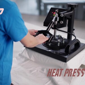 May include: A black heat press machine with a digital control panel and a sturdy metal handle, positioned over a white fabric surface. The device features a flat heating plate, vertical support column, and tension springs for even pressure application.