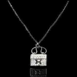 Puede incluir: Un collar plateado con un colgante en forma de peque&ntilde;o bolso. El colgante est&aacute; incrustado con cristales transparentes y presenta la letra "H" en el centro. La cadena es delicada y el fondo es negro.