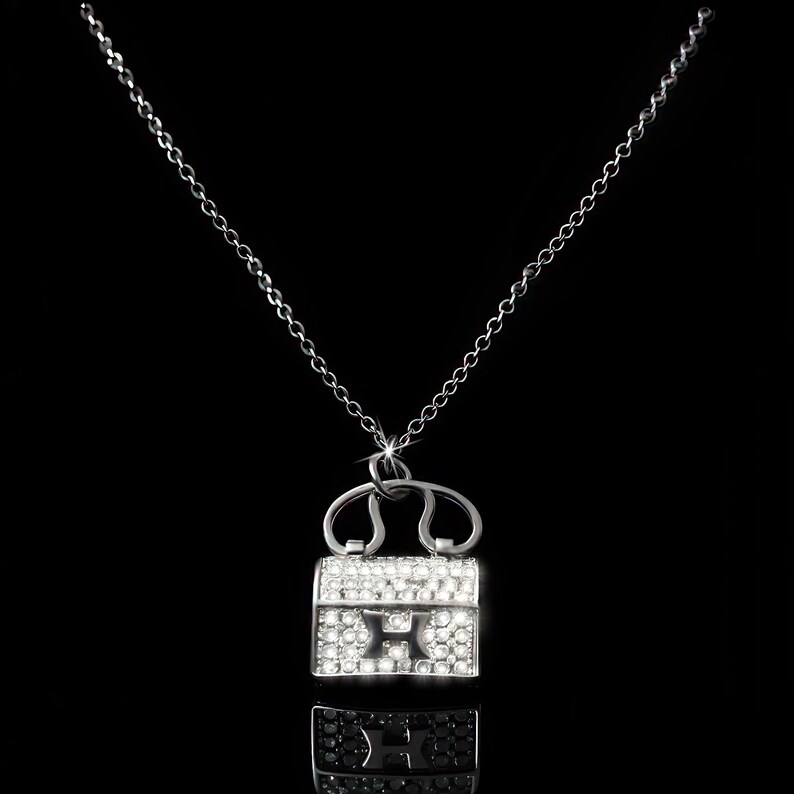 Puede incluir: Un collar plateado con un colgante en forma de peque&ntilde;o bolso. El colgante est&aacute; incrustado con cristales transparentes y presenta la letra "H" en el centro. La cadena es delicada y el fondo es negro.