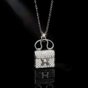 Puede incluir: Collar plateado con un colgante de bolso peque&ntilde;o incrustado con cristales brillantes. El colgante presenta una "H" en el centro y cuelga de una fina cadena. El fondo negro resalta los detalles del colgante.