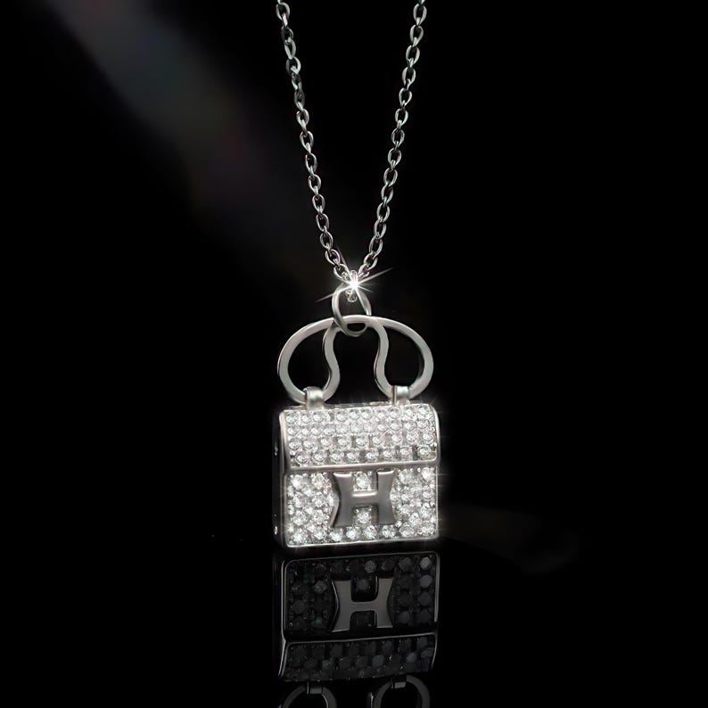 Puede incluir: Collar plateado con un colgante de bolso peque&ntilde;o incrustado con cristales brillantes. El colgante presenta una "H" en el centro y cuelga de una fina cadena. El fondo negro resalta los detalles del colgante.