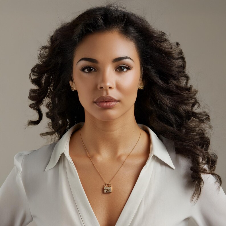 Puede incluir: Una mujer con un collar de oro con un colgante. El colgante es un dise&ntilde;o peque&ntilde;o y rectangular con detalles intrincados. Tambi&eacute;n lleva peque&ntilde;os pendientes de oro. Lleva una camisa blanca.