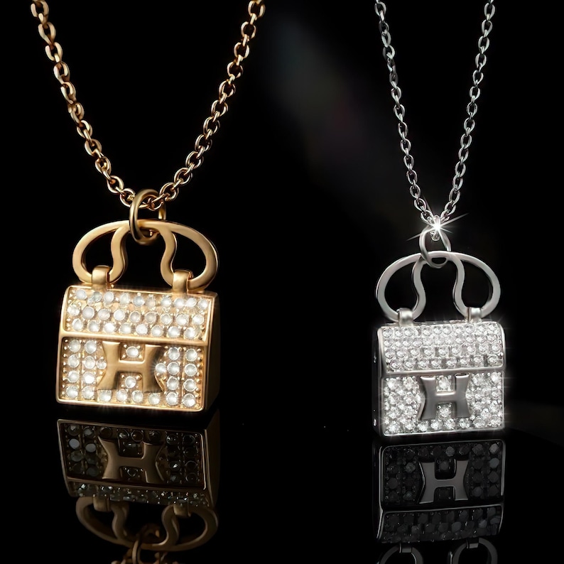 Puede incluir: Dos collares con colgantes de bolso. Uno es dorado, el otro plateado. Ambos colgantes est&aacute;n incrustados con peque&ntilde;os cristales transparentes y presentan un dise&ntilde;o con la letra "H". Los collares cuelgan sobre un fondo negro.