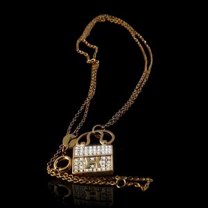 Puede incluir: Collar dorado con un colgante en forma de bolso. El colgante est&aacute; adornado con peque&ntilde;as piedras preciosas transparentes y la letra "H". La cadena es fina y tiene un cierre de mosquet&oacute;n. Fondo negro.