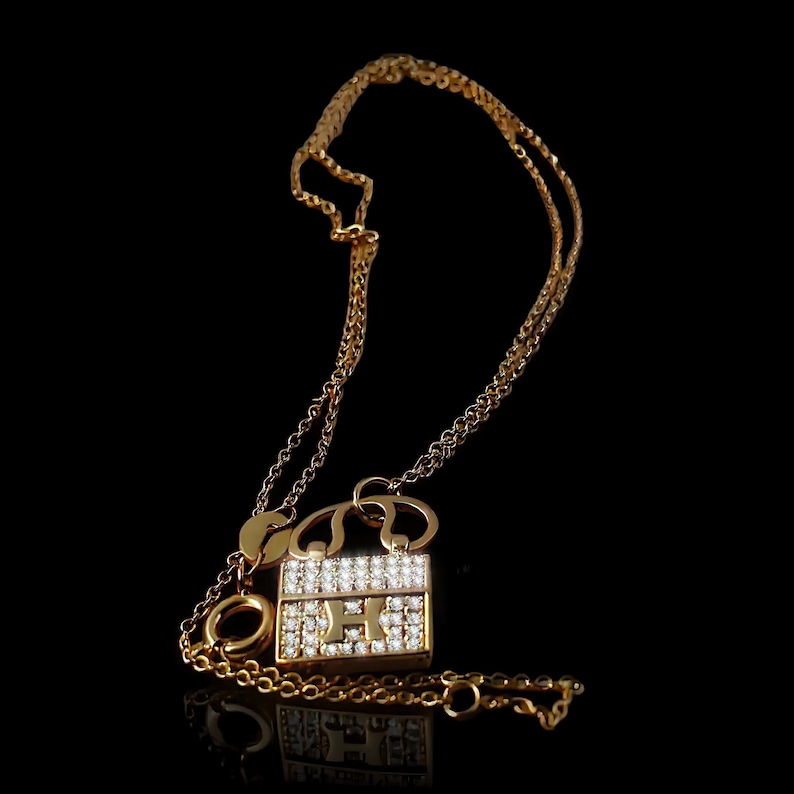 Puede incluir: Collar dorado con un colgante en forma de bolso. El colgante est&aacute; adornado con peque&ntilde;as piedras preciosas transparentes y la letra "H". La cadena es fina y tiene un cierre de mosquet&oacute;n. Fondo negro.