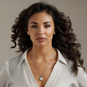 Puede incluir: Un collar de plata con un colgante con dise&ntilde;o de corona y un centro cuadrado, posiblemente con piedras preciosas. El colgante cuelga de una cadena delicada. La mujer lleva el collar con una camisa blanca.