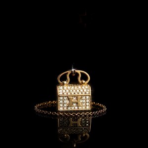 Puede incluir: Un colgante dorado con forma de bolso, adornado con peque&ntilde;as piedras brillantes. El colgante, con una cadena, presenta un dise&ntilde;o con la letra "H". El fondo negro resalta la joya.