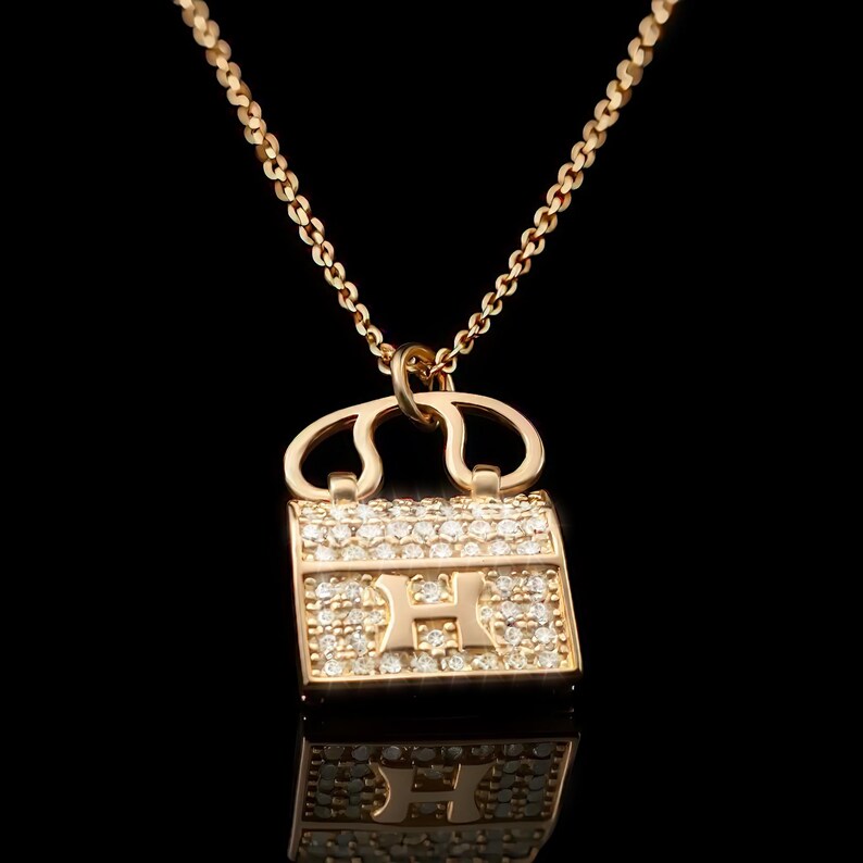 Puede incluir: Collar dorado con un colgante en forma de bolso. El colgante est&aacute; incrustado con peque&ntilde;as piedras preciosas transparentes y tiene un dise&ntilde;o de "H". La cadena es delicada y dorada, con un cierre de mosquet&oacute;n.