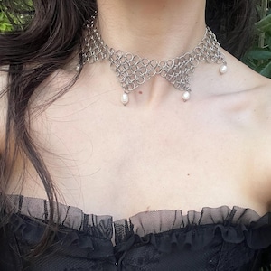 Puede incluir: Un collar gargantilla de cota de malla plateada con tres perlas colgantes. El collar se usa con un top negro. La modelo lleva pendientes de aro y un pendiente con una cruz.