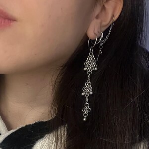 Puede incluir: Pendientes de cota de malla plateados con un diseño colgante. Los pendientes presentan secciones de cota de malla en forma de diamante y pequeñas cuentas plateadas. Una cadena conecta los pendientes a un pequeño aro.