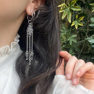 Peut inclure: Boucles d'oreilles en chaîne argentée avec plusieurs brins pendants et de petits accents de boules. Les boucles d'oreilles sont attachées à un cerceau et une chaîne qui remonte l'oreille. Les bijoux sont portés avec un chemisier blanc et des cheveux foncés.