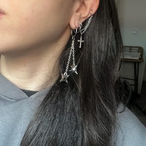 Puede incluir: Pendientes plateados con múltiples aros, un colgante de cruz y dijes en forma de estrella. Los pendientes están conectados por delicadas cadenas, creando un efecto de capas. Diseño moderno y atrevido.