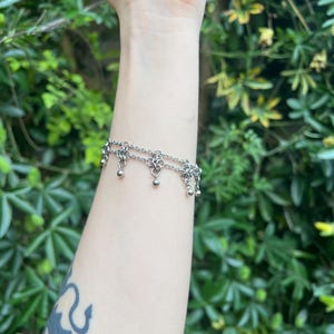 Könnte beinhalten: Ein silbernes Armband mit zwei Kettengliedern und kleinen, baumelnden Kugelanhängern. Das Armband wird an einem Arm mit einem schwarzen Tattoo getragen. Der Hintergrund ist ein grüner Busch.