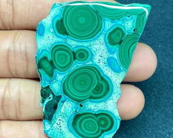 May include: A polished malachite stone with concentric circular patterns in shades of green and teal. The stone has a rough, irregular edge and a smooth, glossy surface. The patterns resemble swirling eyes.