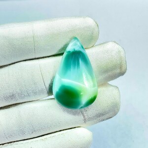 May include: A teardrop-shaped gemstone with a gradient of colors, transitioning from a deep green at the base to a light turquoise at the top. The polished stone has a smooth, glossy surface, and is held in a white glove.