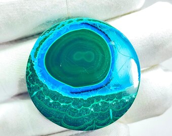 May include: A round, polished gemstone with concentric patterns of green, teal, and blue. The gemstone has a glossy finish and appears to be a natural mineral. The stone is held by a gloved hand.