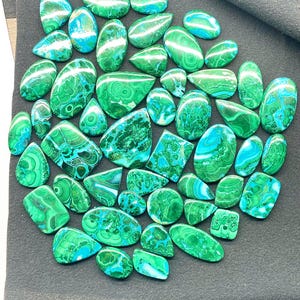 May include: A collection of polished malachite gemstones in various shapes, including ovals and teardrops. The stones display a vibrant green color with swirling patterns and patches of blue-green hues. The gemstones are arranged on a dark gray surface.