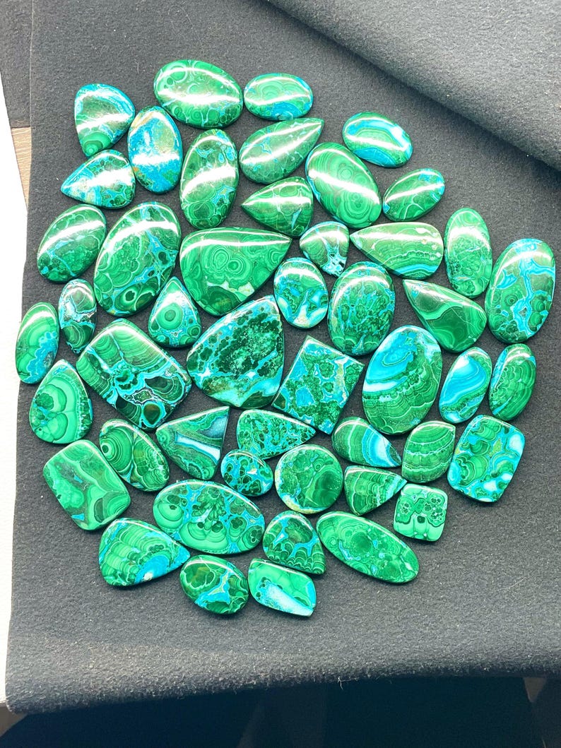 May include: A collection of polished malachite gemstones in various shapes, including ovals and teardrops. The stones display a vibrant green color with swirling patterns and patches of blue-green hues. The gemstones are arranged on a dark gray surface.