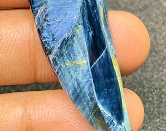 Könnte beinhalten: Ein polierter Edelstein mit einem auffälligen blau-goldenen Muster. Der Stein hat eine glatte, gebogene Oberfläche und eine spitze Spitze. Die Farben verschmelzen und erzeugen ein einzigartiges und auffälliges Aussehen. Der Edelstein wird zwischen den Fingern gehalten.