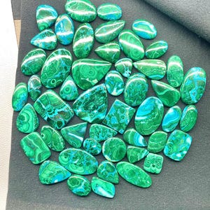 May include: A collection of polished malachite gemstones in various shapes and sizes. The stones display a vibrant green color with swirling patterns and bands of lighter green and blue. The gemstones are arranged on a dark gray surface.