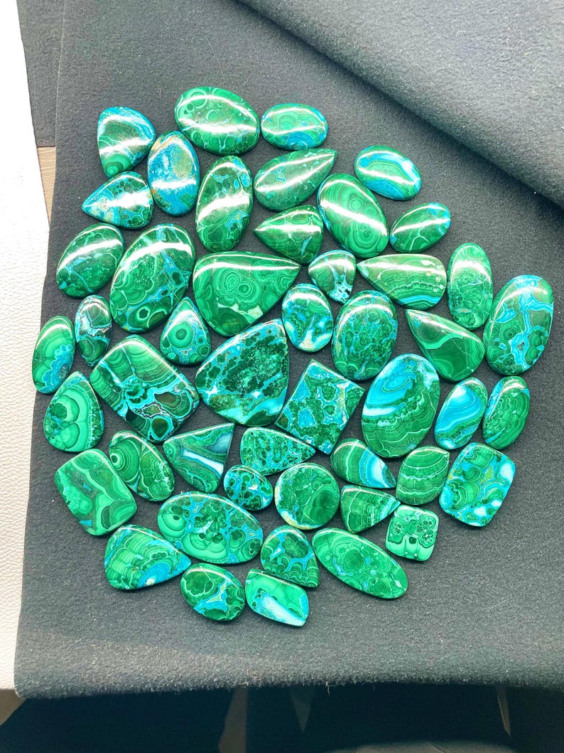 May include: A collection of polished malachite gemstones in various shapes and sizes. The stones display a vibrant green color with swirling patterns and bands of lighter green and blue. The gemstones are arranged on a dark gray surface.
