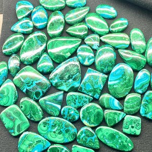 May include: A collection of polished malachite gemstones in various shapes, including ovals, teardrops, and squares. The stones display a vibrant mix of green and blue hues with unique swirling patterns. The gemstones are arranged on a dark surface.