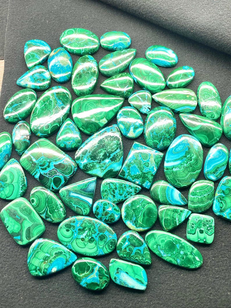 May include: A collection of polished malachite gemstones in various shapes, including ovals, teardrops, and squares. The stones display a vibrant mix of green and blue hues with unique swirling patterns. The gemstones are arranged on a dark surface.