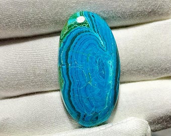 May include: An oval-shaped gemstone with a vibrant blue and green color scheme. The stone features a layered pattern with dark blue and black bands, creating a unique, natural design. The gemstone is set against a white background.