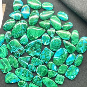 May include: A collection of polished malachite gemstones in various shapes, including ovals, teardrops, and squares. The stones display a vibrant green color with swirling patterns and patches of turquoise blue. The gemstones are arranged on a dark gray surface.