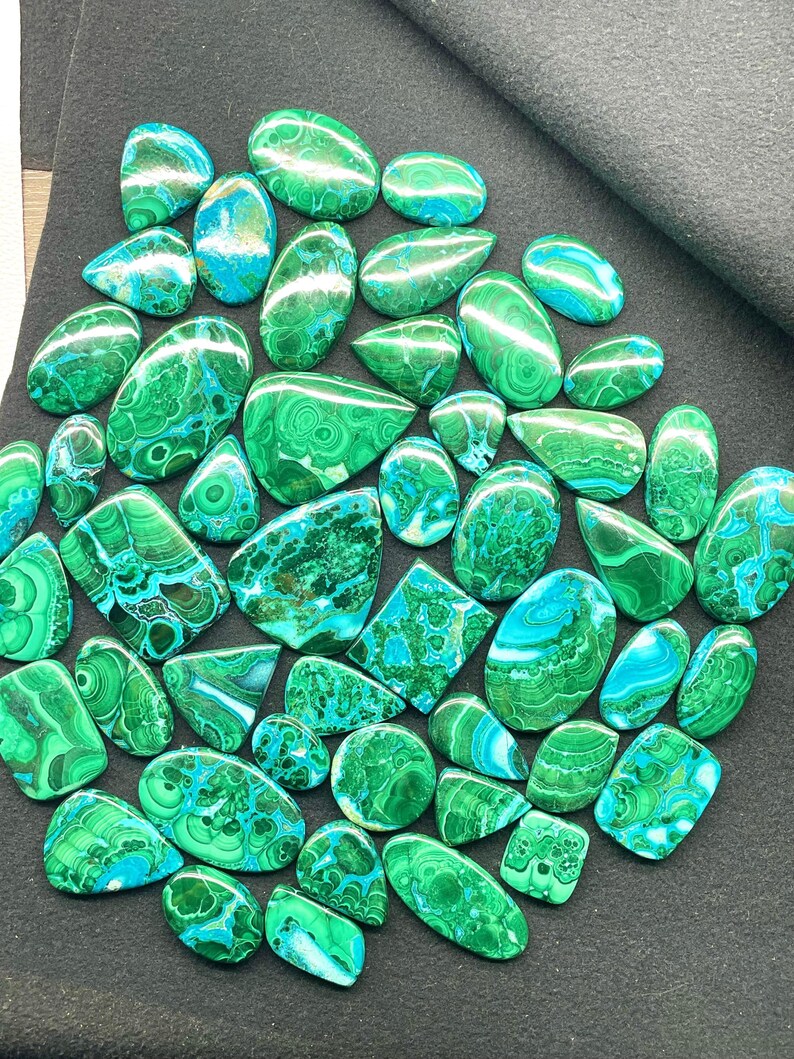 May include: A collection of polished malachite gemstones in various shapes, including ovals, teardrops, and squares. The stones display a vibrant green color with swirling patterns and patches of turquoise blue. The gemstones are arranged on a dark gray surface.