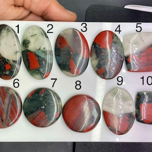 May include: Ten polished gemstones with varied patterns are displayed. The stones feature red, black, white, and green hues. Shapes include oval, round, and square. The gemstones are presented on a white surface.