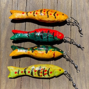 Puede incluir: Cuatro señuelos de pesca de madera pintados a mano en colores vibrantes: amarillo, rojo, verde y naranja. Cada señuelo presenta marcas negras y un accesorio giratorio de metal. Los señuelos están dispuestos sobre una superficie de madera envejecida.