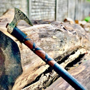 May include: A handcrafted Viking axe with a decorative, etched blade. The axe features a wooden handle with leather wrapping and metal accents. The blade has an intricate design, and the handle is wrapped in black leather. The axe is displayed on a weathered wooden log.
