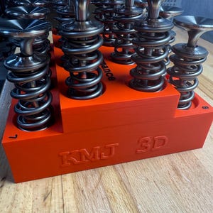 May include: An array of silver-colored valve springs and components are arranged on a bright orange base. The base has the text "KMJ 3D" embossed on the side. The components are for automotive or engine applications.