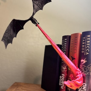 May include: A black dragon bookend with outstretched wings and a fiery breath, positioned between several books. The dragon's breath is a gradient of red and yellow. The books have titles like "Fourth Wing" and "Onyx Storm".