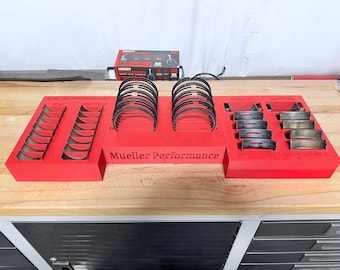 May include: A red Mueller Performance parts organizer with engine components. The organizer has sections labeled "TOP" and "BOTTOM" and holds various metal rings and bearings. A box of Hardy brand parts is visible in the background.