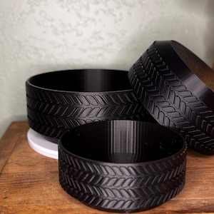 May include: Three black, round containers with a tire tread design. The containers are of varying sizes and are arranged on a wooden surface. The largest container is in the background, with two smaller ones in front.