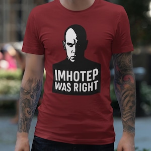 以下が含まれることがあります： 鮮やかなカーディナルレッドの半袖Tシャツ。厳しい表情をした禿頭の人物の白黒グラフィックプリントが施されている。人物の下には、黒い背景に太字の白い大文字で「IMHOTEP WAS RIGHT」というテキストが配置されている。