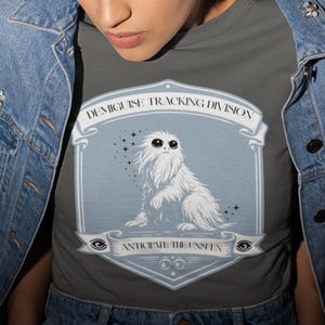 May include: A gray t-shirt featuring a white, fluffy creature illustration with the text "DEMIGUISE TRACKING DIVISION" and "ANTICIPATE THE UNSEEN". The shirt is worn with a blue denim jacket.