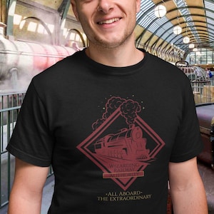 Può includere: Una t-shirt nera a maniche corte con una grafica rosso scuro di una locomotiva a vapore all'interno di una cornice a rombo. Il testo recita Wizarding Railway Authority e All Aboard The Extraordinary, con fumo che sale dal treno.