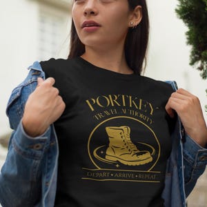 May include: Black t-shirt with a gold graphic featuring a boot inside a circle, the words "PORTKEY TRAVEL AUTHORITY" and "DEPART. ARRIVE. REPEAT." The shirt is worn by a person in a denim jacket.