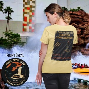 Op de afbeelding: Boterkleurig T-shirt met de tekst "SURVIVED THE RAPTOR CONTAINMENT" en een dinosaurusafbeelding. De voorkant toont "FINAL DROP CLUB" met coördinaten. Het shirt wordt gedragen voor een waterattractie.